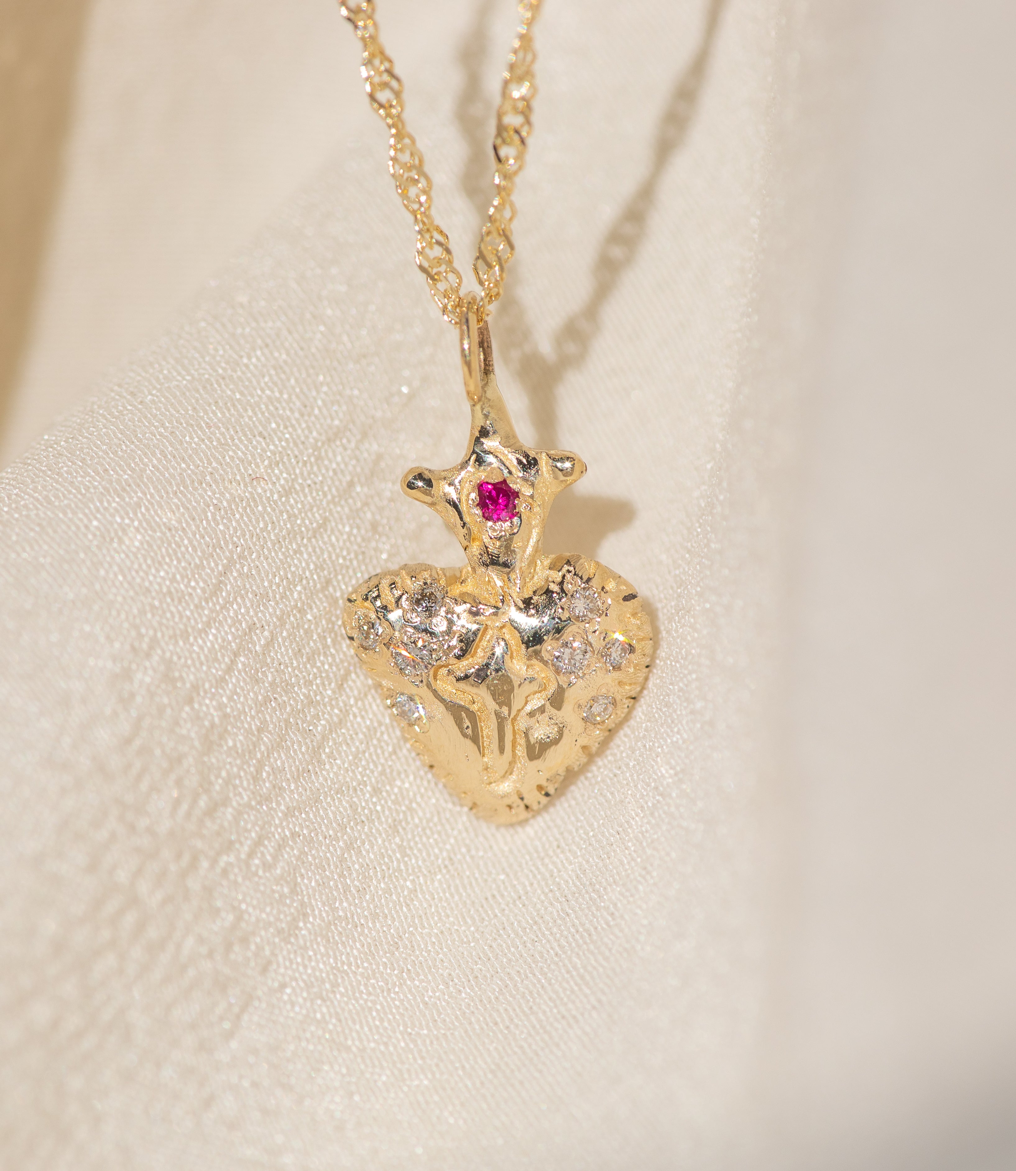 Gold necklace with a heart-shaped pendant featuring a red gemstone on a beige background
