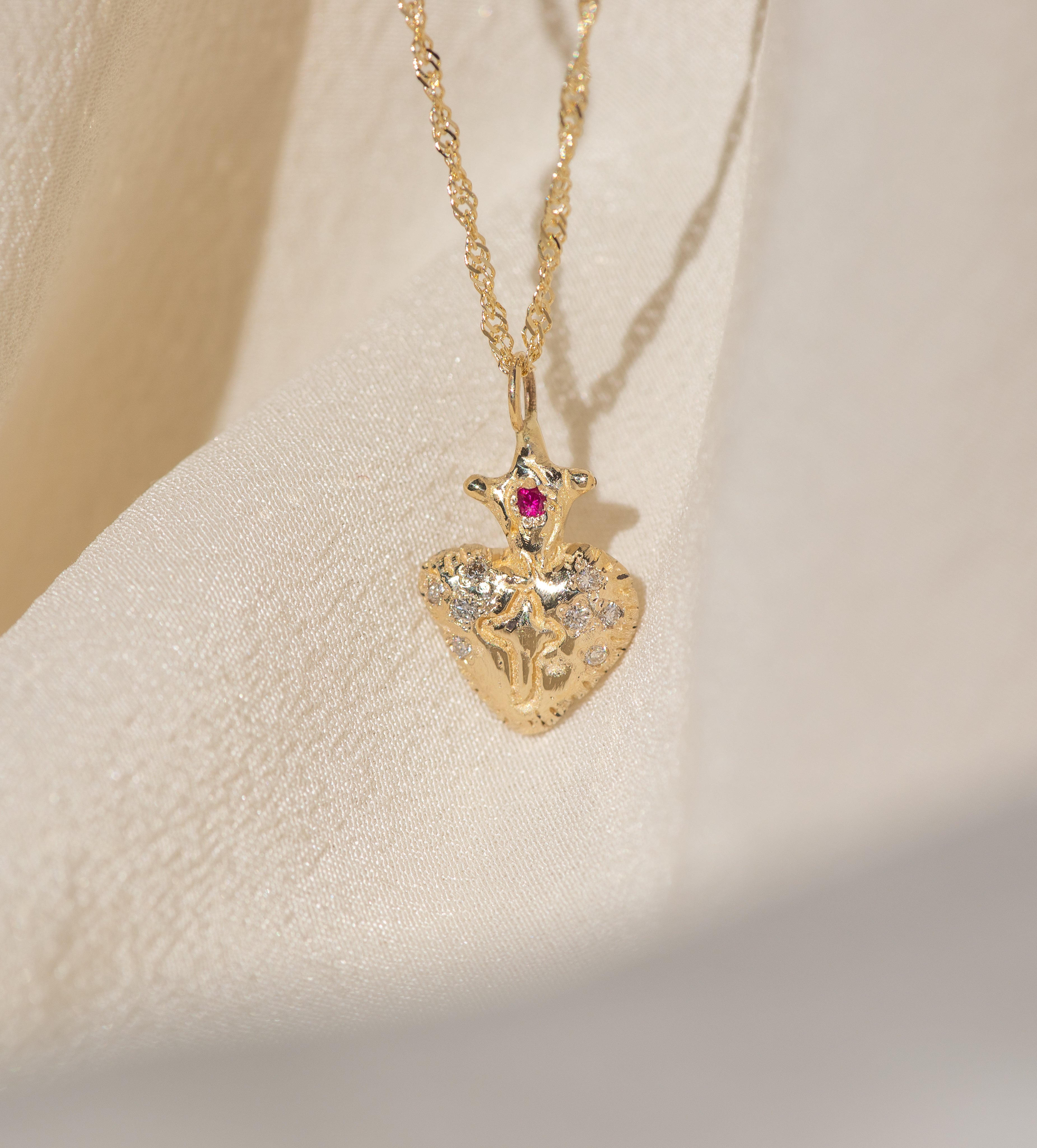 Gold necklace with a heart-shaped pendant featuring a red gemstone on a beige background