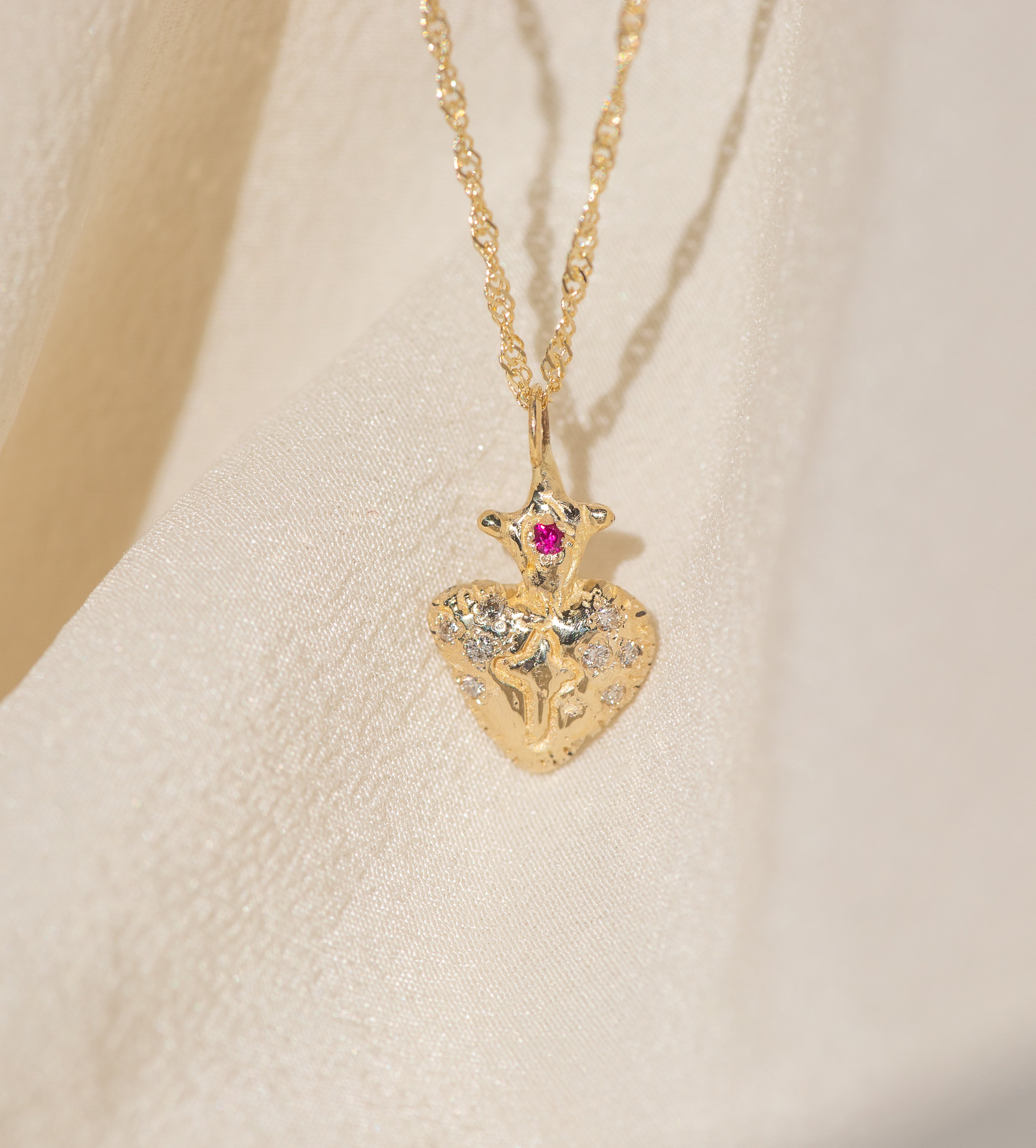Gold necklace with a heart-shaped pendant featuring a red gemstone on a beige background